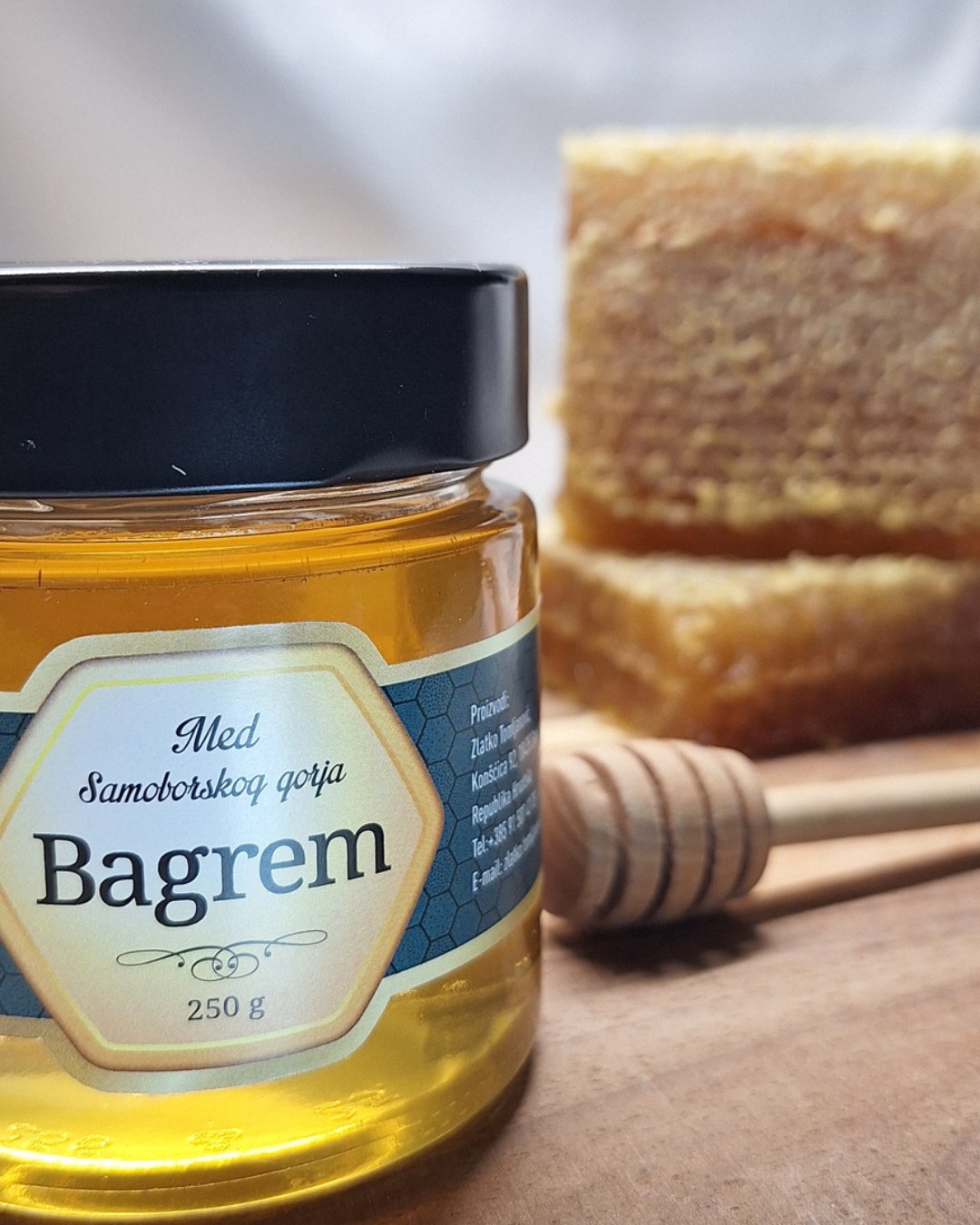 Jar of honey with 'Bagrem' label, honeycomb, and honey dipper on wooden surface